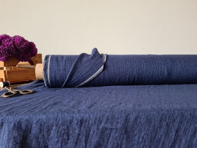 May include: A roll of dark blue fabric with a textured appearance. The fabric is partially unrolled, revealing a lighter edge. A wooden crate with purple flowers and a pair of scissors are in the background.