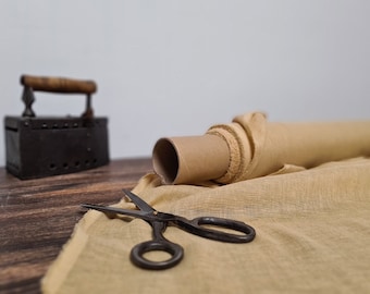 Può includere: Un rotolo di tessuto color marrone chiaro su un tubo di cartone, un paio di forbici in metallo nero e un ferro da stiro vintage in metallo nero con manico in legno. Il tessuto ha un aspetto intrecciato e testurizzato ed è steso sulla superficie.