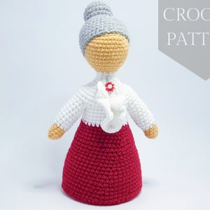 Puede incluir: Patrón de crochet para una muñeca con un moño gris, una blusa blanca y una falda roja. La muñeca tiene una cara de crochet con un botón rojo para la nariz.