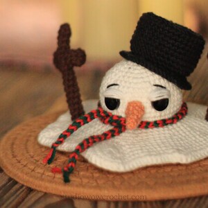 May include: A crocheted snowman with a black top hat, red and green scarf, and brown stick arms. The snowman is melting on a brown surface.