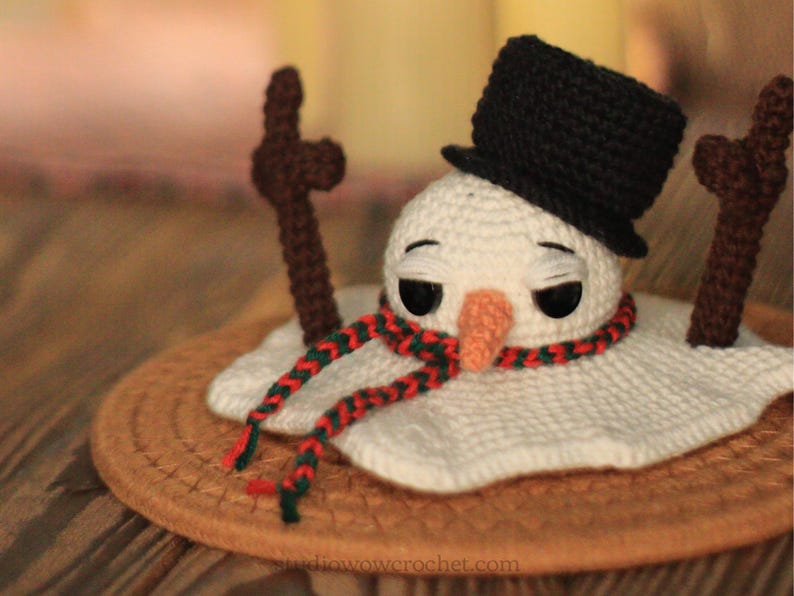 May include: A crocheted snowman with a black top hat, red and green scarf, and brown stick arms. The snowman is melting on a brown surface.
