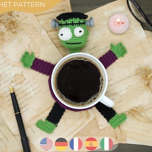 Op de afbeelding: Een gehaakte Frankenstein-monster koffiekopjeshouder met groene, zwarte, paarse en grijze details. De armen en benen van het monster strekken zich uit de beker, die gevuld is met koffie. De afbeelding bevat een pen, kaarsen en tekst.