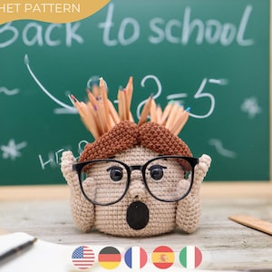 Puede incluir: Un portalápices de ganchillo con forma de cara sorprendida, con gafas negras y bigote marrón. El portalápices está lleno de lápices y colocado frente a una pizarra con la inscripción "Back to school".