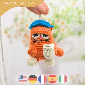 Peut inclure: Chat orange au crochet avec un chapeau bleu, tenant une tasse blanche. Le chat a des moustaches noires et un nez rose. Le texte "CROCHET PATTERN" est en haut à gauche. Les pieds du chat sont blancs.