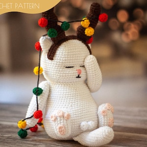 May include: White crocheted cat wearing a brown crocheted reindeer antler headband with colourful string lights. The cat is sitting on a wooden surface.