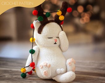 May include: White crocheted cat wearing a brown crocheted reindeer antler headband with colorful string lights. The cat is sitting on a wooden surface.