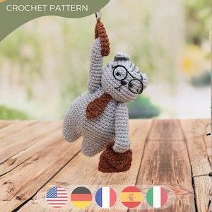 May include: A crocheted grey cat wearing glasses, a brown tie, and carrying a brown briefcase. The cat is hanging from a brown loop. The text "CROCHET PATTERN" is at the top.