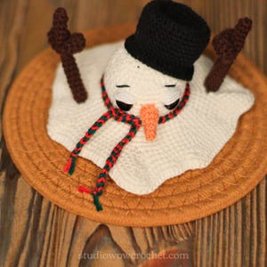 May include: A crocheted snowman with a black hat and a red and green scarf, sitting on a brown woven mat.