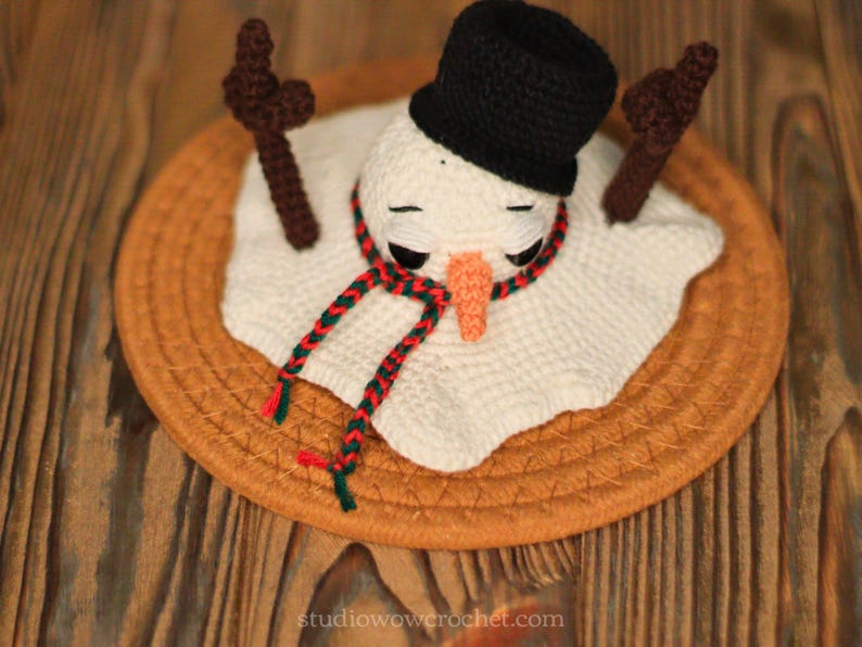 May include: A crocheted snowman with a black hat and a red and green scarf, sitting on a brown woven mat.