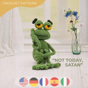 Puede incluir: Una rana verde tejida a ganchillo con grandes ojos amarillos y negros y pequeños mechones negros. La rana está levantando los brazos y tiene el texto "NOT TODAY, SATAN" junto a ella. La imagen también incluye el texto "CROCHET PATTERN".