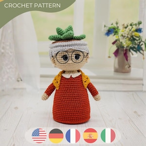 Puede incluir: Una muñeca de ganchillo con cara color canela, gafas redondas negras y cabello gris rematado con una planta suculenta verde. La muñeca viste un vestido color óxido con cuello blanco y un chal texturizado amarillo. El texto CROCHET PATTERN es visible.