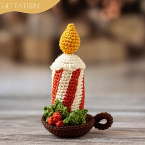 Puede incluir: Una vela de ganchillo con un cuerpo a rayas rojas y blancas, una llama amarilla y hojas verdes con bayas rojas en la base. La vela está sentada sobre una superficie marrón.