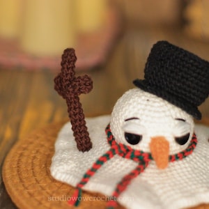 May include: A crocheted snowman wearing a black top hat and a red and green scarf. The snowman is melting and has brown sticks for arms. The snowman is sitting on a brown woven mat.