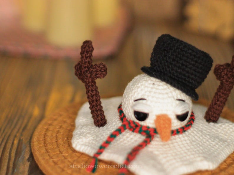 May include: A crocheted snowman wearing a black top hat and a red and green scarf. The snowman is melting and has brown sticks for arms. The snowman is sitting on a brown woven mat.