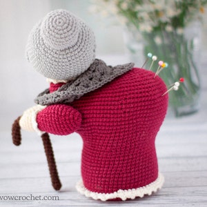 May include: A crocheted pincushion shaped like an elderly woman wearing a red dress, grey shawl, and a grey hat. The pincushion has a brown walking stick and several colorful pins stuck in its back.