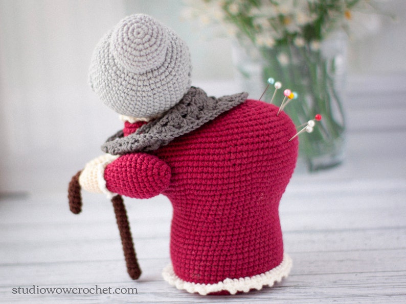May include: A crocheted pincushion shaped like an elderly woman wearing a red dress, grey shawl, and a grey hat. The pincushion has a brown walking stick and several colorful pins stuck in its back.