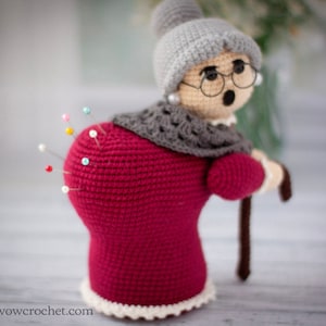 May include: A crocheted pincushion shaped like a woman wearing a red dress, grey shawl, and a grey hat. The pincushion has a white lace trim and is holding a brown walking stick.  There are colorful pins stuck into the back of the pincushion.