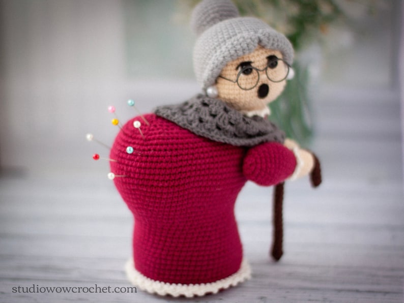 May include: A crocheted pincushion shaped like a woman wearing a red dress, grey shawl, and a grey hat. The pincushion has a white lace trim and is holding a brown walking stick.  There are colorful pins stuck into the back of the pincushion.