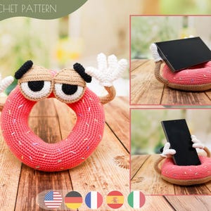 Könnte beinhalten: Ein gehäkelter Donut-Handyhalter mit einem Cartoon-Gesicht, Armen und Händen. Der Ständer ist rosa mit weißen Streuseln und einem braunen Rand. Der Text "CROCHET PATTERN" steht oben. Der Ständer ist für ein Handy oder Tablet.
