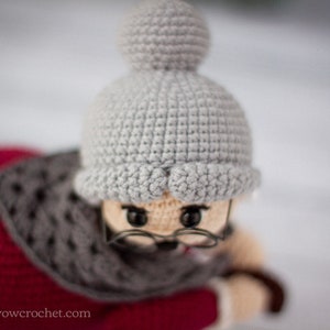 May include: A crocheted doll wearing a gray hat with a pom-pom and a red and gray scarf. The doll has brown eyes and is wearing glasses.