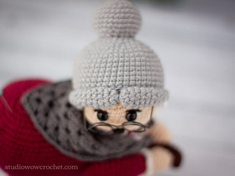May include: A crocheted doll wearing a gray hat with a pom-pom and a red and gray scarf. The doll has brown eyes and is wearing glasses.