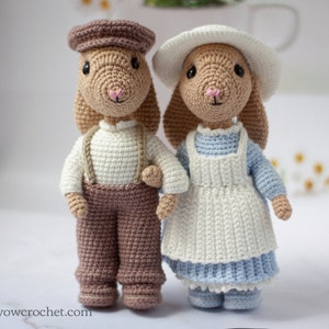 Puede incluir: Dos muñecas de conejo de crochet, una con una gorra marrón y pantalones marrones, la otra con un sombrero blanco y un vestido azul con un delantal blanco.