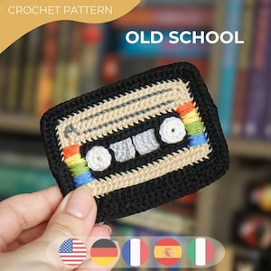 Op de afbeelding: Een gehaakt cassettebandontwerp met de woorden "OLD SCHOOL" in het wit. De cassetteband is zwart met beige en grijze details en een regenboogkleurige streep. De woorden "CROCHET PATTERN" staan bovenaan.