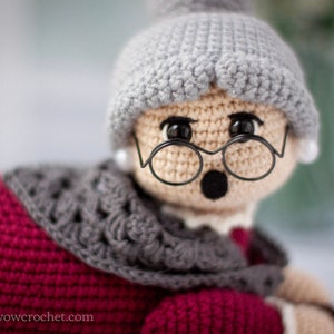 May include: A crocheted doll with gray hair, a red sweater, and a gray scarf. The doll is wearing glasses and has a surprised expression.