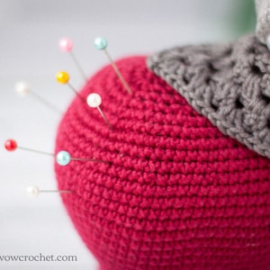 May include: A close-up of a red crocheted pincushion with colorful pins inserted. The pincushion is topped with a gray crocheted piece.