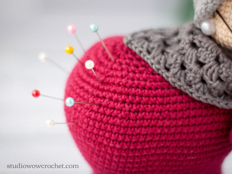 May include: A close-up of a red crocheted pincushion with colorful pins inserted. The pincushion is topped with a gray crocheted piece.