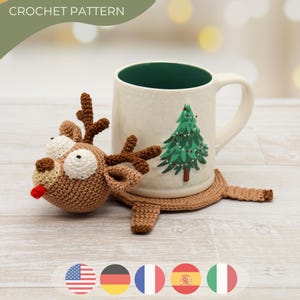May include: A crocheted reindeer coaster with a brown body, white eyes, and a red tongue, paired with a white mug featuring a green Christmas tree design. The text "CROCHET PATTERN" is at the top.