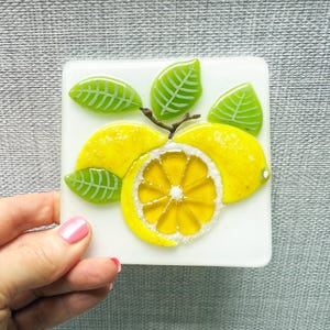 May include: A square, white decorative tile with a lemon design. The design features bright yellow lemons, a sliced lemon, and green leaves. The tile is held by a hand with pink nail polish.