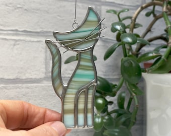 May include: A stained glass cat figurine with a silver frame. The cat is in a sitting position with a long tail and whiskers. The glass is a mix of green, white, and tan colors. The figurine is hanging from a silver wire.