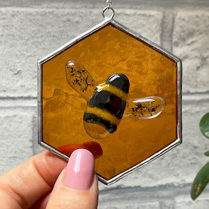 May include: A stained glass bee hanging ornament in a hexagonal frame. The bee is yellow and black with clear wings. The frame is a honey color.
