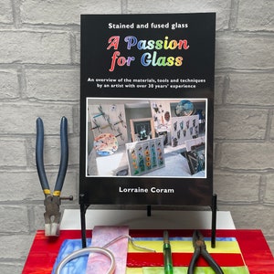 May include: A black book titled "A Passion for Glass" with a colourful title and a photo of stained glass art on the cover. The book is on a black stand and there are glass cutting tools on a table in front of the book.
