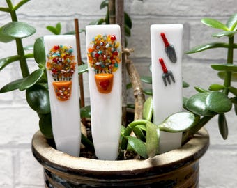 May include: Three white plant markers with colourful designs. Two markers feature a flower pot design with colourful glass beads. The third marker has a trowel and a garden fork with red handles. The markers are in a ceramic pot with green plants.