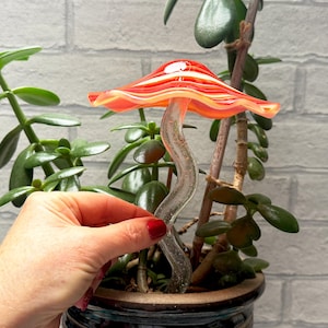 May include: A decorative glass mushroom with a red and orange striped cap and a clear, curved stem. The mushroom is placed in a potted plant with green leaves, against a white brick wall.