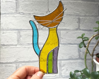 May include: A stained glass cat suncatcher with a yellow, orange, green, blue, and purple design. The cat is hanging from a wire.