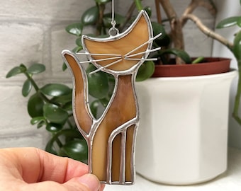 May include: A stained glass cat suncatcher with a brown body and silver accents. The cat is sitting up with its tail curled around its body.