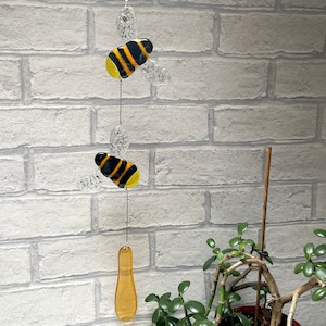 May include: A glass bee wind chime with two black and yellow bees and a yellow glass teardrop hanging from a string. The wind chime is hanging from a white brick wall.
