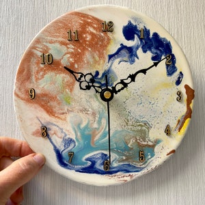 May include: A round, white clock with a blue, brown, and yellow marbled design. The clock face has black hands and gold numbers.