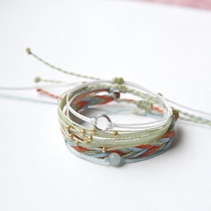 May include: A stack of colorful friendship bracelets with a silver charm and gold beads. The bracelets are made of woven string and feature a light blue, green, and orange color scheme.