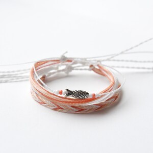 May include: A multi-strand bracelet with a woven design in shades of peach and white. The bracelet features a small silver fish charm.