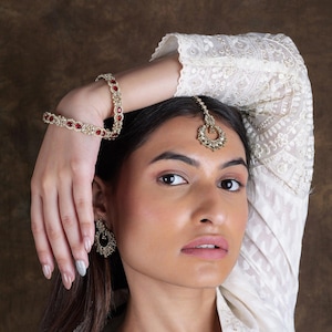 May include: A woman wearing a white embroidered top with a gold and red jeweled headpiece, earrings, and bracelets.