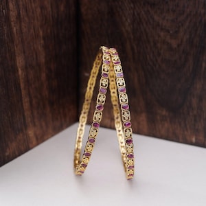 Può includere: Due braccialetti d'oro con un motivo ripetuto di piccole pietre preziose rosse ovali e minuscole pietre trasparenti. I braccialetti sono appoggiati su una superficie bianca con uno sfondo in legno marrone scuro.