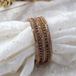 Op de afbeelding: Een set van vier gouden armbanden met rode en heldere strass steentjes. De armbanden zijn op elkaar gestapeld en liggen op een witte stoffen achtergrond.