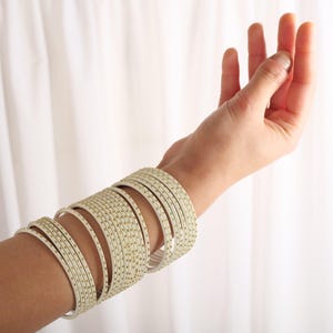 Może przedstawiać: Stos białych bransoletek z złotymi akcentami. Bransoletki są noszone na nadgarstku.