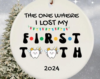 May include: White ceramic Christmas ornament with the text "The One Where I Lost My First Tooth" and the year 2024. The text is in black with a red, green, yellow, and blue string of lights above the word "First". Two cartoon tooth fairies with wings and wands are on either side of the word "Tooth".