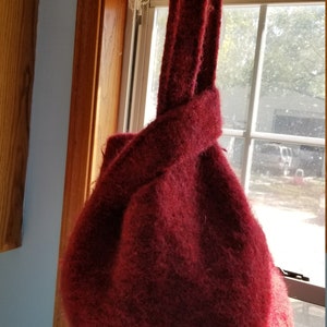 May include: A deep red knitted tote bag with a knotted handle. The bag is hanging from a wooden rod.