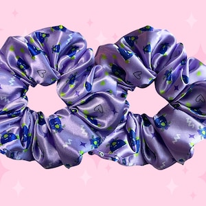 Puede incluir: Dos coleteros de satén lavanda con un estampado repetido de figuras alienígenas azules, puntos verdes, zigzags blancos y estrellas y corazones morados. Los coleteros están fruncidos, creando un aspecto voluminoso.